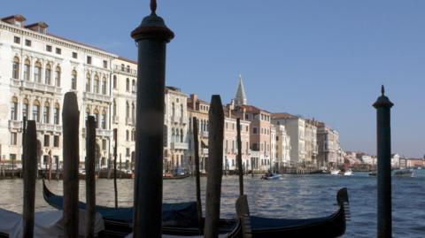 A waterfront in Venice with wooden posts and gondolas in the foreground and a row of buildings and more boats in the background.