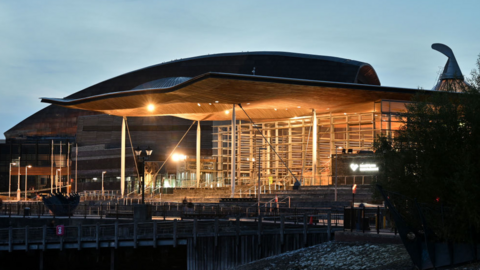 An exterior picture of the Senedd building with its lights on in the early evening