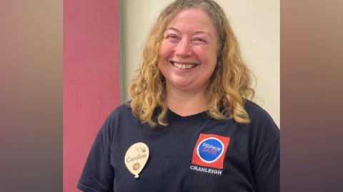 A blonde woman wearing a dark navy shirt, with a logo that reads Repair Cafe Cranleigh. She is also wearing a badge that reads Caroline.