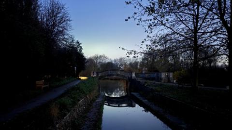 A canal at night with reflections in the water of a bridge over the water. The sky is a dark blue with a slight hint of yellow on the horizon as the sun sets. There is a path on each side of the water and trees are in silhouette.