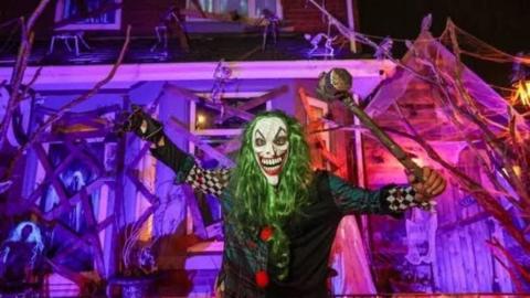 A man is wearing a scary white clown mask, long green hair, a tartan, checkered and black clown costume, one black glove with long silver nails and is holding a mallet. Behind his is an end terraced house, covered in skeletons, wooden planks, cobwebs and has a huge wooden extension. The decorations are lit by purple and blue lights.