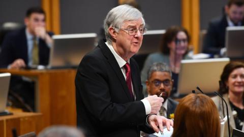 Mark Drakeford addressing the Senedd surrounded by politicians from the Welsh parliament. He's wearing a black suit and red tie.
