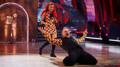 Chris McCausland doing a knee slide on the strictly dance floor with Diane Buswell behind him. Both in 60s inspired costumes