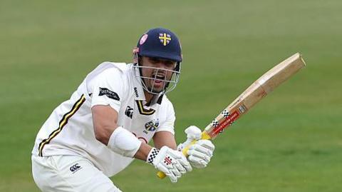 Left-hander Emilio Gay, wearing his navy blue Durham helmet, watches after playing a shot into the legside with his bat in the air 