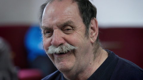 Michael Edwards smiling at the camera. He has dark hair and a white moustache. He is wearing a dark blue top. The background is blurred but he appears to be in a room.