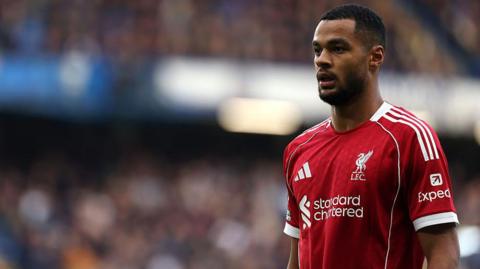 Cody Gakpo, wearing Liverpool's red home kit, standing still during a match with the crowd a blur in the background