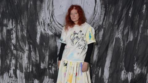 Delaine La Bas is standing in front of a black and white artwork. She is wearing a paint-stained effect skirt and T-shirt, the shirt shows an image of a heart with "THE HEART" written on it in capital letters.