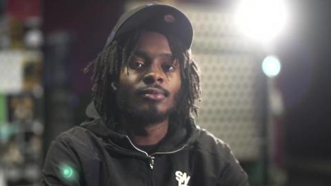 Malachi Saunders, a young man with short dreadlocks wearing a black cap and black hoodie, sitting in front of bright studio lights