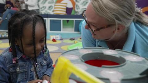 A woman wearing glasses on the right who works for the parenting network, she is playing with a young girl on the floor while parents can be seen in the background talking