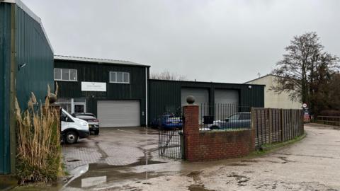 A green building in an L-shape with a car park in front within gates on a rainy day.