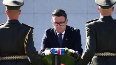 Andy Burnham wearing a dark suit leans over a wreath, he is seen between two soldiers