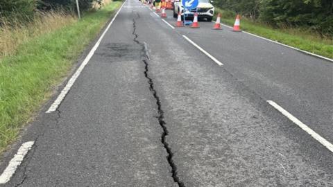 A road with a long crack in the middle of the left-hand lane. There are grass verges on both sides of the road. Orange and white cones care in the distance on the right side of the road, with cars parked behind them.