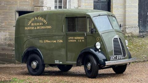An olive green-coloured original Morris JE van is parked up on gravel in front of a stone building. It has 'Smith & Grieves Builders Painters & Decorators' painted on the side, along with 'Tel: Livingstone 3586' and 'Upper Norwood. London. S E 19'.  