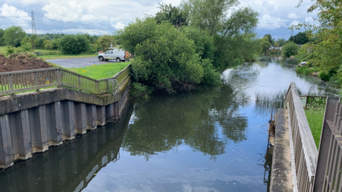 The view from a footbridge over the River Soar with a van parked on the left bank near to foliage hanging over the water