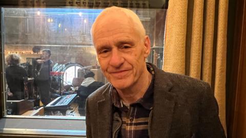 Steve Macarthur is standing inside the Cheese and Grain in front of a window looking through to a recording studio. He is wearing a jacket and is smiling at the camera.