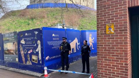 Two police officers standing in a walkway. There are cones in front of them with police cordon tape. On the left of the officers is a blue wall with advertising for Norwich Castle. On the right, there is a brick wall. Behind the this is a grassy hill with Norwich Castle on top. 