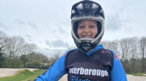 Leah Pearson, in a light and dark blue top saying Peterborough Phantoms. She is wearing a helmet and smiling broadly. Behind her are undulating concerte tracks and leafless trees against a grey sky.