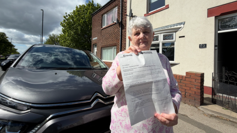 Joan Everett, who is wearing a pink floral dress, stands in front of her and her home while holding up a letter. The letter has been issued by Sunderland Council, which confirms the road will close and she must park at the rear of her home is she is to continue using her car.