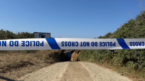 Blue and white police tape cordons off a road in Sark during a fire. The writing on the tape says "police do not cross". The writing is upside down.