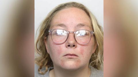 Police custody image of Stacey Sharples who has shoulder length blond hair and pale pink rimmed glasses and wearing a grey t-shirt.