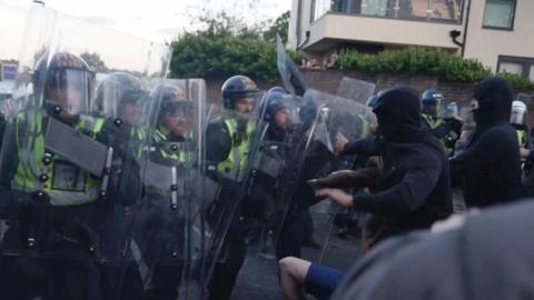 People wearing face coverings and their hoods up push forward towards a line of police officers with riot shields and helmets on