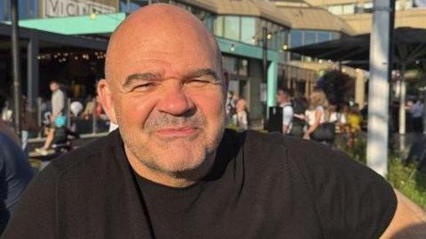 A head and shoulders shot of Paul Hughes. He has greying short stubble, is wearing a dark T-shirt and is about to smile. He is outside and behind him is a shopping centre with people walking around. 