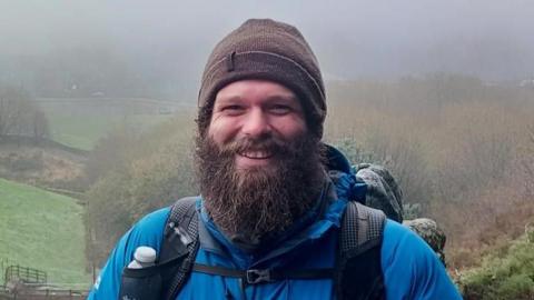 Owen Charnley, who is wearing a blue coat, brown hat, bags on his shoulders, and has a large brown beard, smiles while standing overlooking a misty field shrouded by trees
