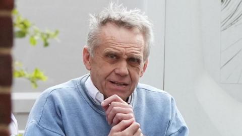 An elderly man wearing a blue jumper over a white suit shirt. He has white hair and is standing in front of a white background with red bricks in the foreground to the left.