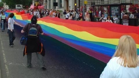 Demonstrators carrying a rainbow flag down a city street. Spectators and cameramen can be seen at the side of the street their are marching past.