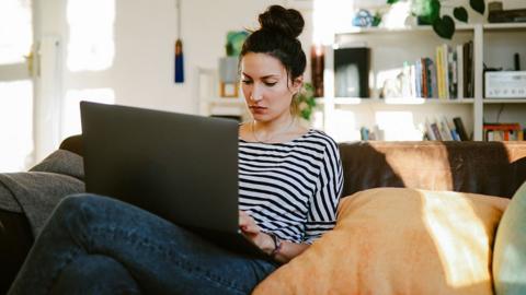 A woman wearing a black and white striped jumper sits on a brown sofa using a laptop, with shelving and other living room designators in the background