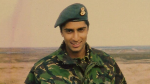 Flight Lieutenant Rakesh Chauhan smiles at the camera.  He is wearing green camouflage fatigues and a blue beret