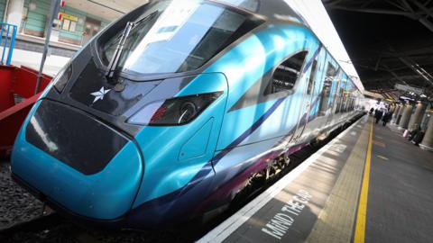 A modern blue train with a pointed front pulled up on tracks alongside a platform with a sign for Manchester Airport seen behind