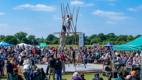 An acrobatic performance of two people standing on bamboo sticks with people watching them. 