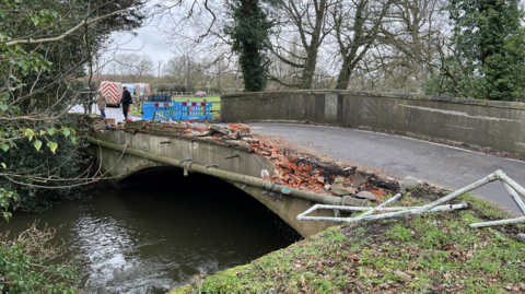 A general view picture of the bridge, which has seen its side closest to the camera totally flattened, with metal railings bent out of place and bricks scattered nearby. 