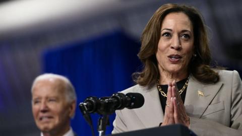 Kamala Harris with her hands held in promise, with Joe Biden in the background