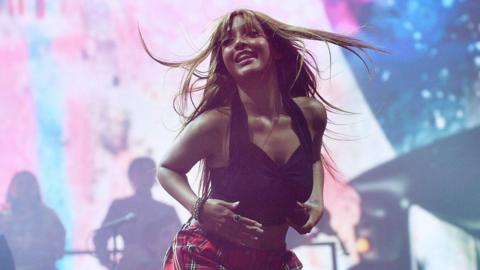 A female singer on stage, wearing a black vest top, tartan trousers and holding a mic.