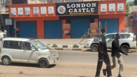 Two men walk past a shuttered restaurant and parked cars on a quite street in Juba during the daytime. The restaurant is called 'London Castle' and features a Union Jack flag.