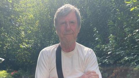 Leslie Midwinder, a man with short hair brushed to the side, is pictured looking at the camera. He is stood outside in front of some green trees and bushes and wearing a white T-shirt.