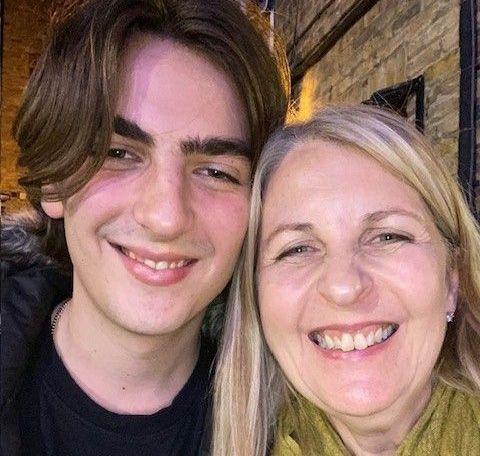 Alex Harpum (left) smiles at the camera with his mouth slightly open, showing a glimpse of his teeth. His hair is centre parted and flops over his right eye slightly. He has his head leaned against that of his mother Anne Jacques who also grins with her teeth showing in this picture and squints her eyes, while wearing a green scarf like a pashmina. 