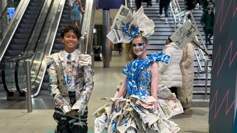 Dominic and Aoife are wearing sustainable outfits they made, with an assortment of newspaper clippings, plastic bags, train tickets and tape. They are standing and smiling at the camera. 
