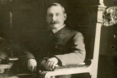 John is sitting in a grand wooden chair. He has a moustache and is looking at the camera. It is a black and white photograph. He is wearing a dark suit jacket and white collar.