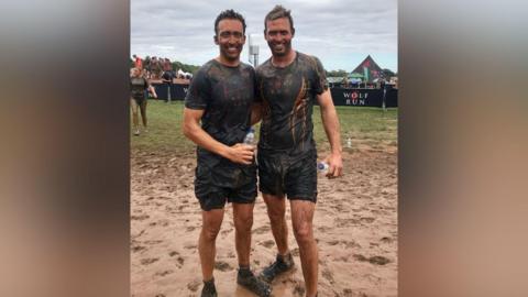 Two brothers standing arm in arm in a very muddy field that is saturated in mud. They are wearing black T-shirts with writing on that says "wolf run". they are covered in mud from head to toe and are smiling.