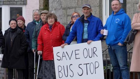 A group of Mousehole residents gathered in the village centre with one holding a placard saying 'Save Our Bus Stop'