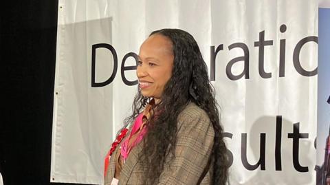 Nicola Jenkins wearing a checked court and a pink lanyard- standing on a podium in front of a white and black board