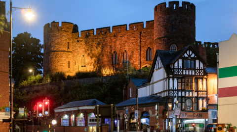 The wall of a castle in the evening with round turrets at either end and a collection of illuminated buildings below, including a black and white timbered building