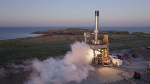 A cylindirical silver rocket without a nose cone on the launch pad at a peninsula on an island in Shetland. White vapour is emerging from its thrusters as it prepares for launch. In the background is the sea.