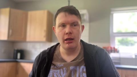 Mid-shot James Patton in a kitchen. He has brown hair, a brown t-shirt and a black cardigan.