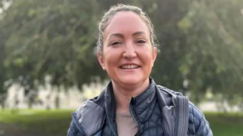 A woman with tied-back brown hair is wearing a grey coat and is standing in front of trees and greenery.