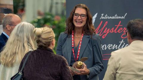 A woman laughs as she talks to a small crowd of people. She's holding an award, seems like she just won it and is proudly talking to some friends 