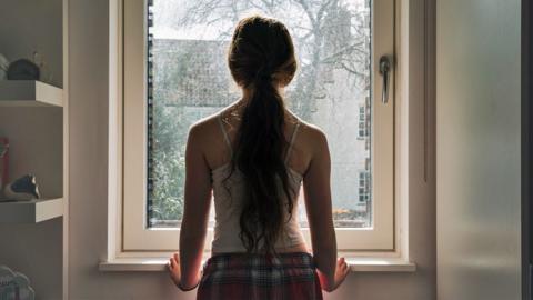 Stock image of a girl with long hair tied in a ponytail stands next to a window and looks outside, wearing a vest and checked pyjama bottoms.